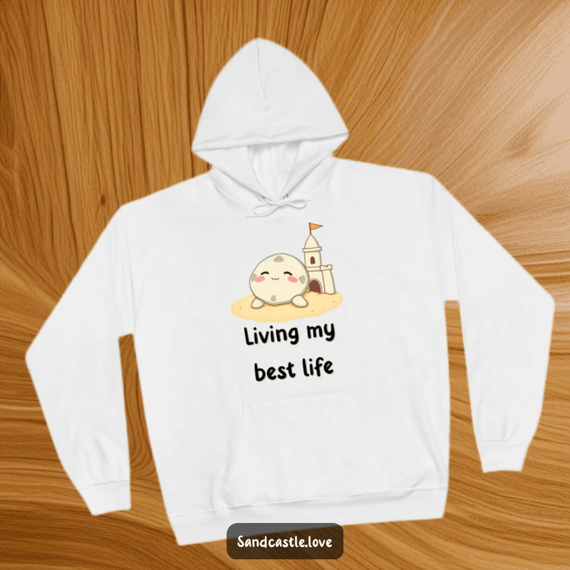 Funny sand dollar hoodie, a contented character resting on warm sand beside a sandcastle, comfortable humorous apparel.